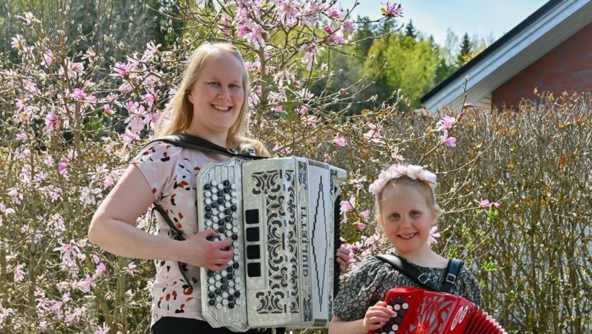 Aikuinen ja lapsi seisovat harmonikat syissä. Taustalla on vaaleanpunaisin kukin kukkiva puu.