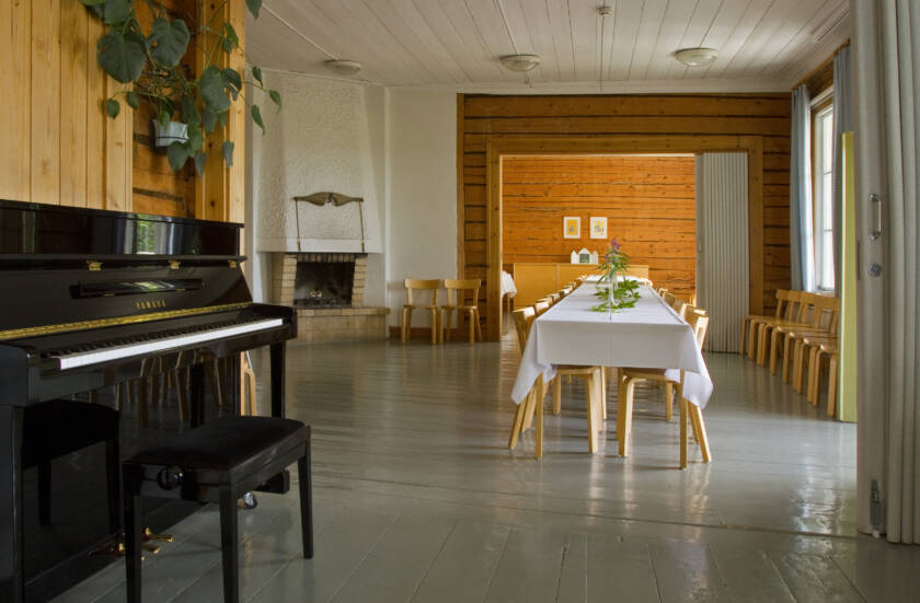 Tomt rum med ett svart piano, ett långt vitt bord och trästolar. I bakgrunden en öppen spis och växter på väggen.