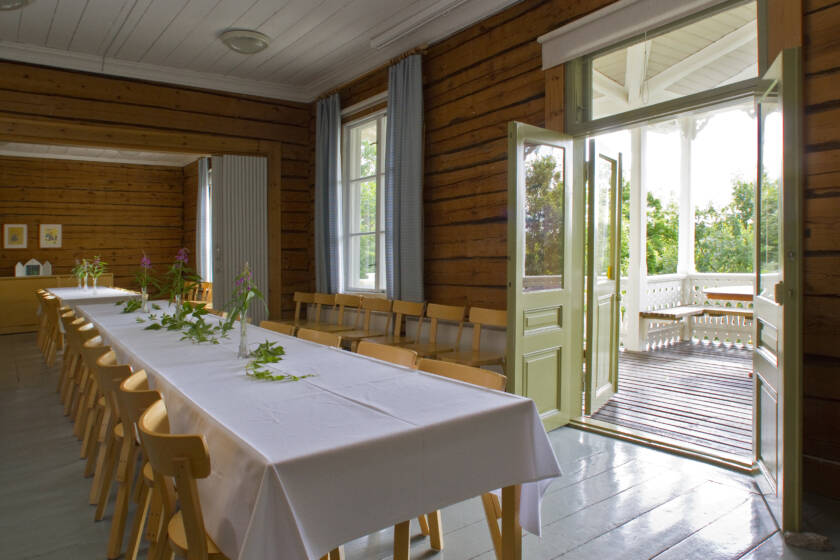 Ett rymligt rum med ett långt bord täckt av en vit duk. Fönstren är öppna, ljus strömmar in och gröna träd syns utanför.