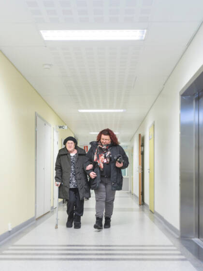 Vapaaehtoinen saattaa vanhusta. Sairaalan käytävä. Kuva Timo Jakone