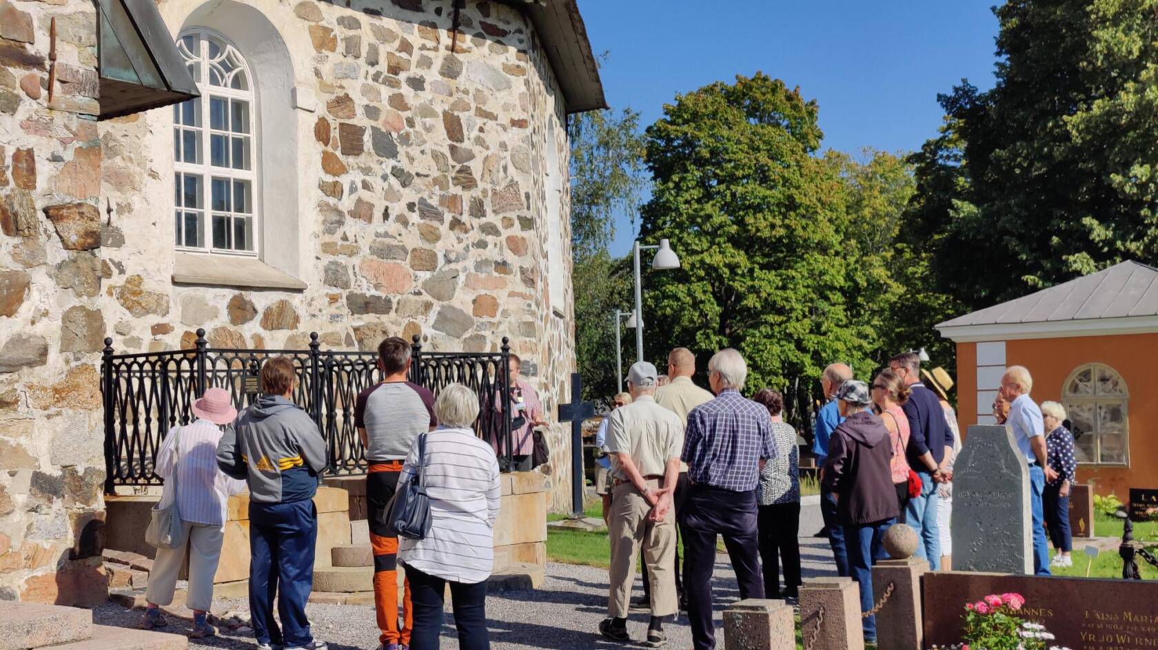 Ihmisiä seisomassa kivikirkon ulkopuolella hautausmaalla.